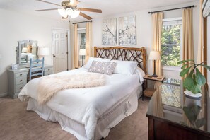 Ein Schlafzimmer mit Bett, Deckenventilator, Kommode, Nachttisch, Lampe, Spiegel und einem Fenster mit Vorhängen.