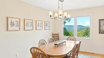A dining area with a wooden table and chairs, a chandelier, framed botanical prints on the wall, and a view of greenery through large windows.