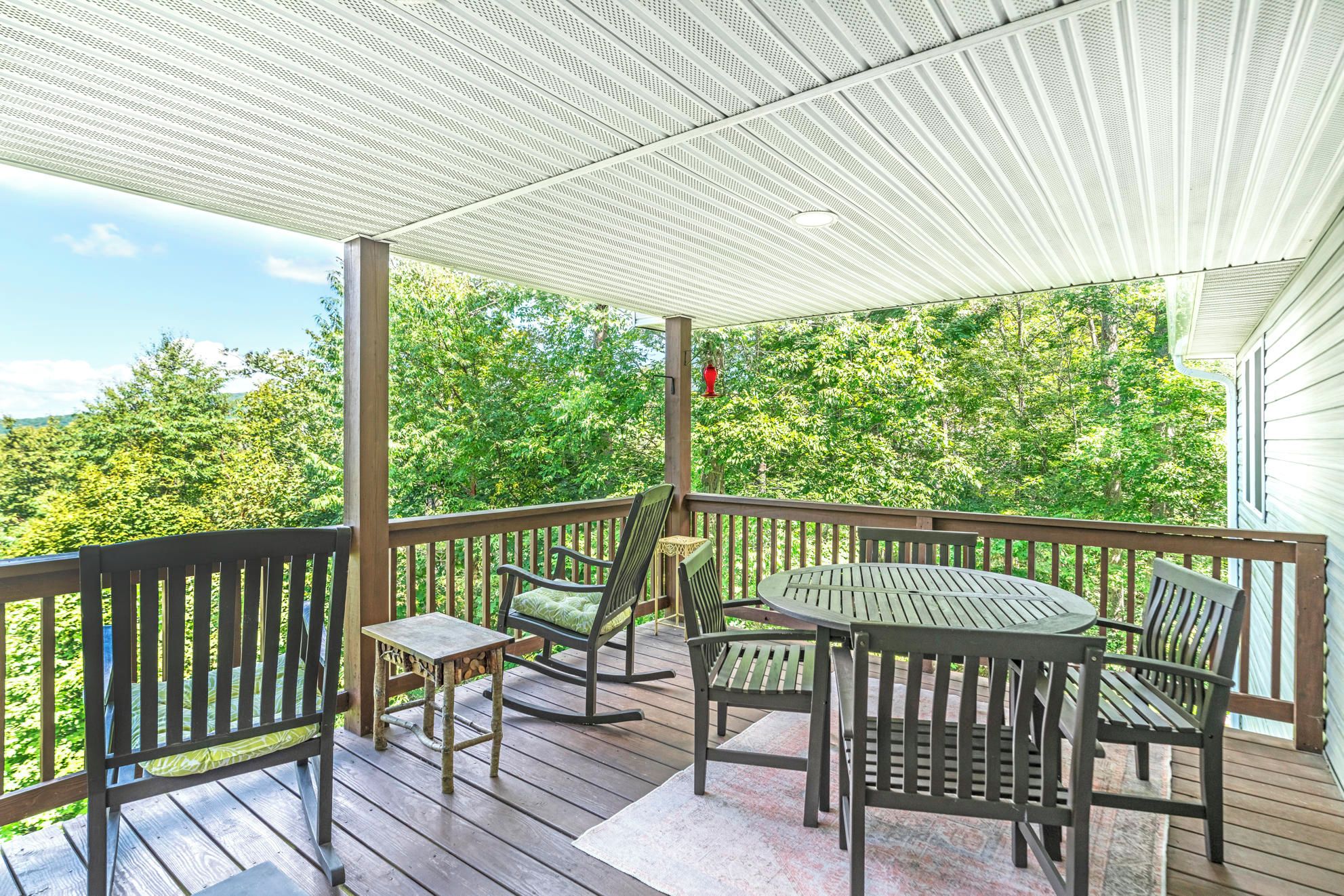 A covered outdoor deck with a table and chairs, surrounded by a wooden railing, overlooking a lush green landscape.