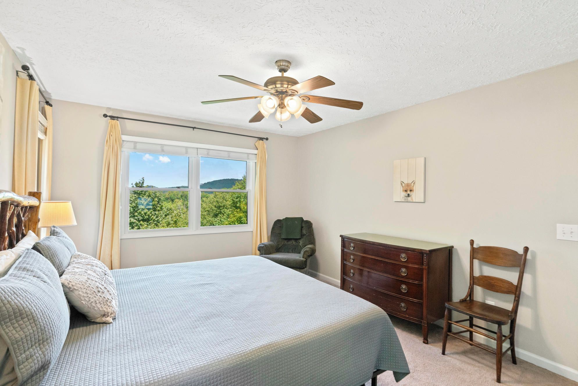 Une chambre à coucher comprenant un lit, une commode, une chaise, un ventilateur de plafond et une fenêtre donnant sur un paysage verdoyant.