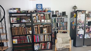 Een kamer met een grote boekenkast vol boeken, een rieten stoel, een klein tafeltje met afstandsbedieningen en een koelkast versierd met foto’s en notities.