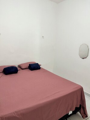 A single bed with a pink bedspread and two dark pillows, a white wall with a small hole, and a white circular object on the wall.