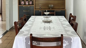 Een eetruimte met een tafel gedekt voor zes personen, omringd door houten stoelen. Een wit kantkleed bedekt de tafel, en erboven hangt een lamp.