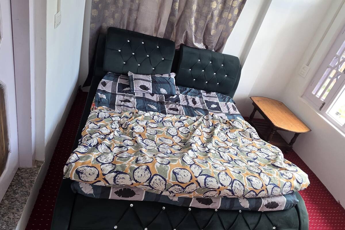 Um quarto com cama, uma mesinha, ventilador de teto e janela com cortinas.