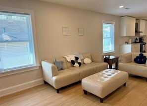 A living room with a beige sofa, an ottoman, and a kitchen area in the background. - New renovation in the heart of Ventnor Heights (Ventnor City)