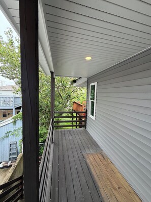 A covered porch with a wooden deck, a railing, and a view of a residential area with trees and a blue fence. - New renovation in the heart of Ventnor Heights (Ventnor City)