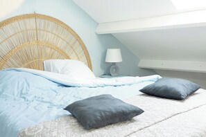 A bedroom with a rattan headboard, a bed with blue bedding, two pillows, a bedside lamp, and a white ceiling. - Casa Rosa (Dinard)