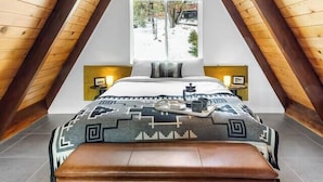 Un dormitorio con techo inclinado, una cama con colcha estampada, un banco y una ventana con vistas a árboles.