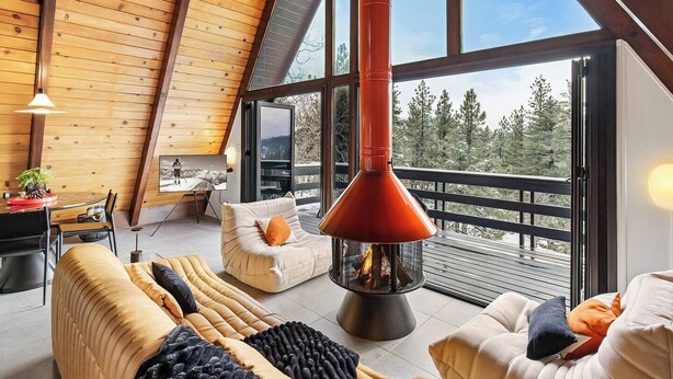 Un interior moderno con techo alto, vigas de madera, chimenea y un gran ventanal con vistas a un bosque.