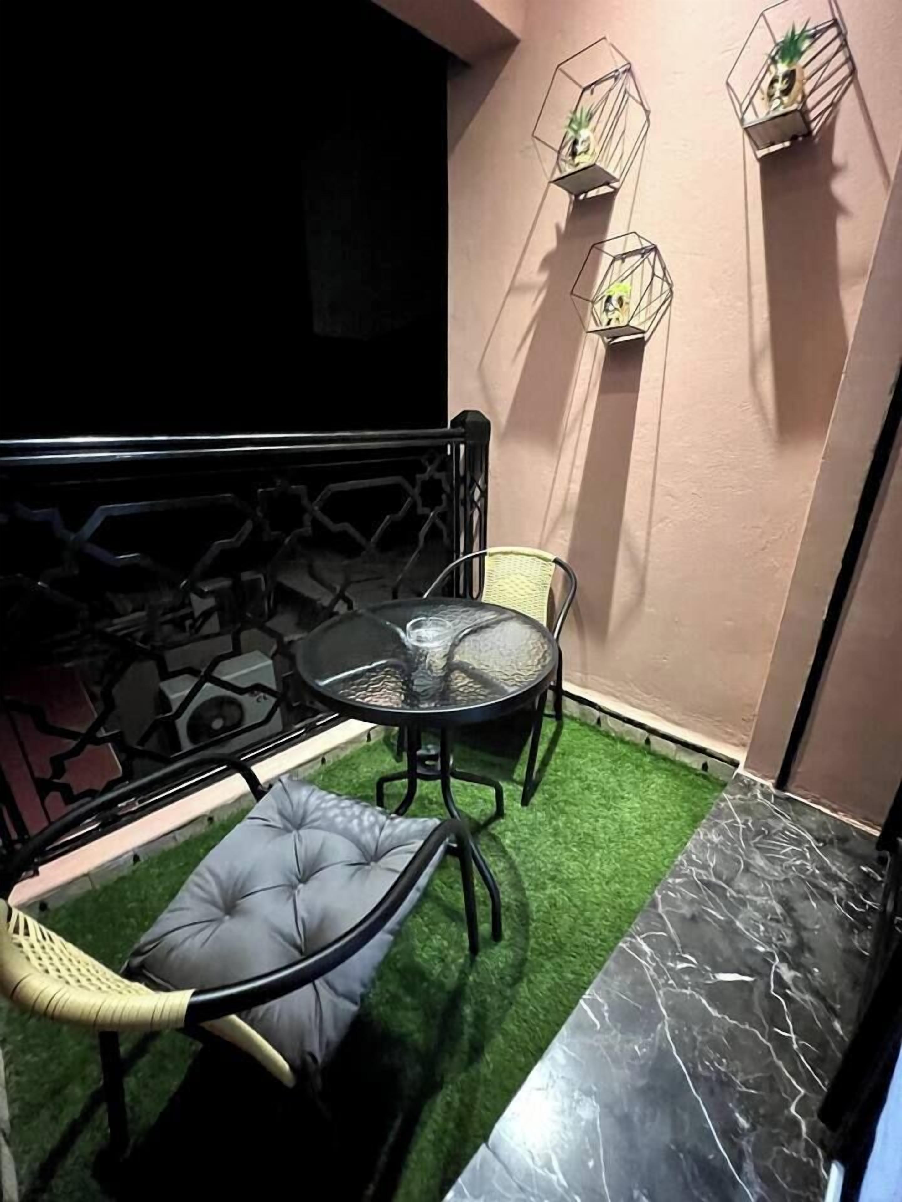 Een klein balkon met een ronde tafel en twee stoelen, een vloer van kunstmatig gras in de kleur groen, een zwarte metalen reling, en drie zeshoekige plantenbakken met beplanting.