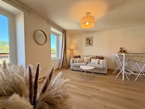 Ein Wohnzimmer mit einer Couch, einem kleinen Tisch, einer Lampe, einem Essbereich mit Stühlen, einem Spiegel und Blick durch ein Fenster.