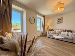 A living room with a sofa, a small round table, a lamp, and a view of a mountain through the window. - Les Coteaux d'Anaïs - Tournon-sur-Rhône (Tournon-sur-Rhône)