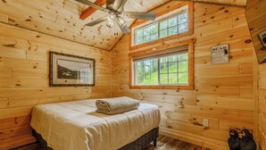 A wooden cabin room with a bed, a ceiling fan, two windows, a framed picture, and a bear sign.