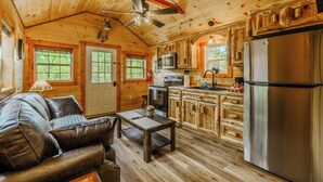 Interior de una cabaña de madera con sofá, mesa de comedor, zona de cocina y ventilador de techo.