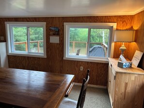 Une pièce avec deux fenêtres, un mur en bois, une table, une chaise, une lampe et un meuble.