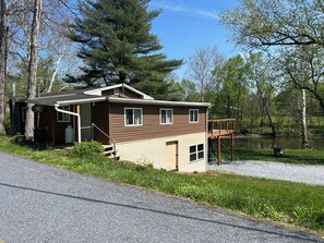 A cabin with a deck, a small porch, and a pond in the background.