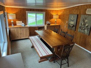 A cabin-style room with a wooden interior, a large dining table, benches, and two lamps.