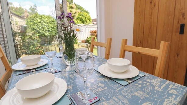 Una mesa de comedor preparada con cuencos blancos, copas de vino y un florero con flores, con vista a follaje exterior.