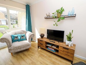 A living room with a sofa, television, wooden cabinet, and a window with curtains. - Camellia Cottage (Wimborne)