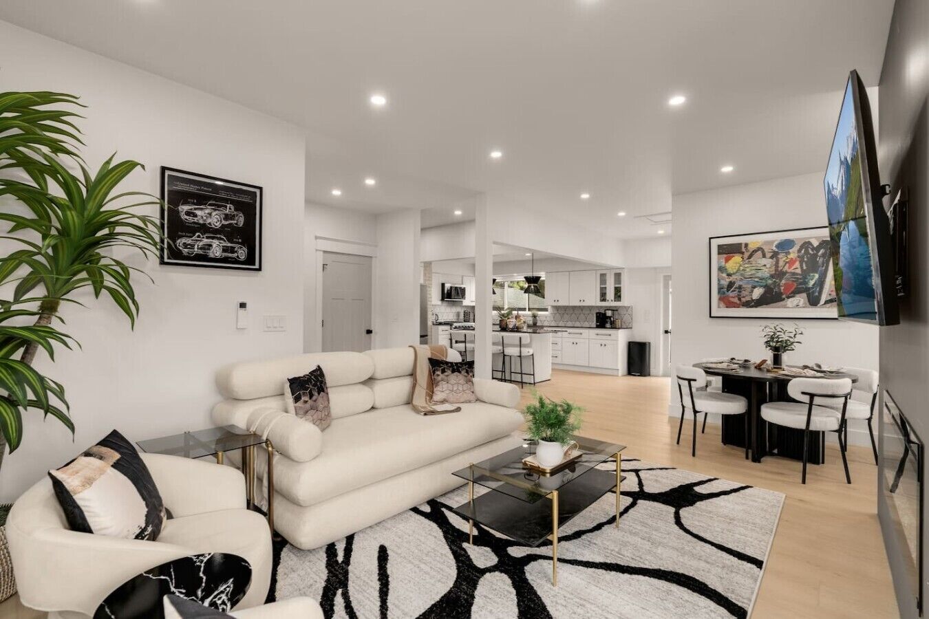 Una sala de estar moderna con un sofá, una mesa de centro y una zona de comedor con sillas. Se observa una cocina al fondo con gabinetes blancos y una mesa de comedor.