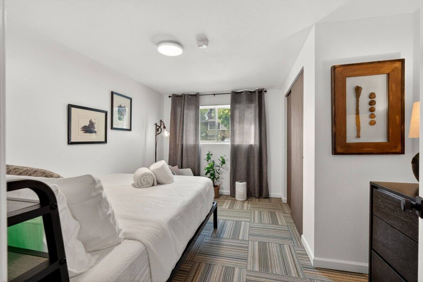 A hotel room with a bed, a desk, a chair, a plant, and a window with curtains.