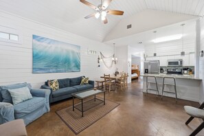 A spacious living area with a large painting, a ceiling fan, a dining table, and a kitchen area with modern appliances. - Pirates Bay 3BR Getaway w/ Lagoon-Style Pool (Port Aransas)