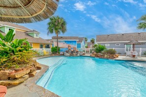 A swimming pool with a rock feature, surrounded by colorful buildings and palm trees. - Pirates Bay 3BR Getaway w/ Lagoon-Style Pool (Port Aransas)