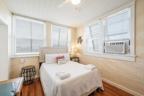 A bedroom with a bed, two bedside tables, a nightstand, a wall-mounted air conditioner, and a window with blinds. - Coastal Studio with Pool and Beach Boardwalk (Port Aransas)