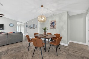 A dining area with a round table, six chairs, a hanging light fixture, and a clock on the wall. - Heated Pool & Hot Tub, Walk to Beach, Fully Equipped. Free Bikes. Grill. (Naples)