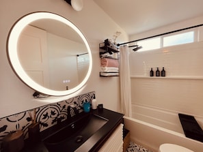 A bathroom with a round mirror, a black sink, a towel rack, and a shower area. - Cozy 3-bedroom cabin in charming North Fork with AC (North Fork)