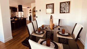 A dining area with a table set for four, surrounded by chairs, with artwork on the walls and a kitchen in the background. - Cozy 3-bedroom cabin in charming North Fork with AC (North Fork)