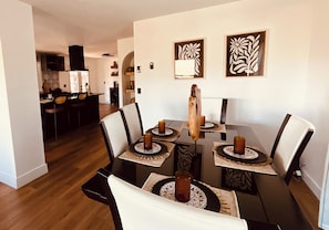 A dining area with a table set for four, surrounded by chairs, with artwork on the walls and a kitchen in the background. - Cozy 3-bedroom cabin in charming North Fork with AC (North Fork)