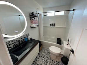 A bathroom with a round mirror, a sink, a toilet, a shower, and a patterned floor. - Cozy 3-bedroom cabin in charming North Fork with AC (North Fork)
