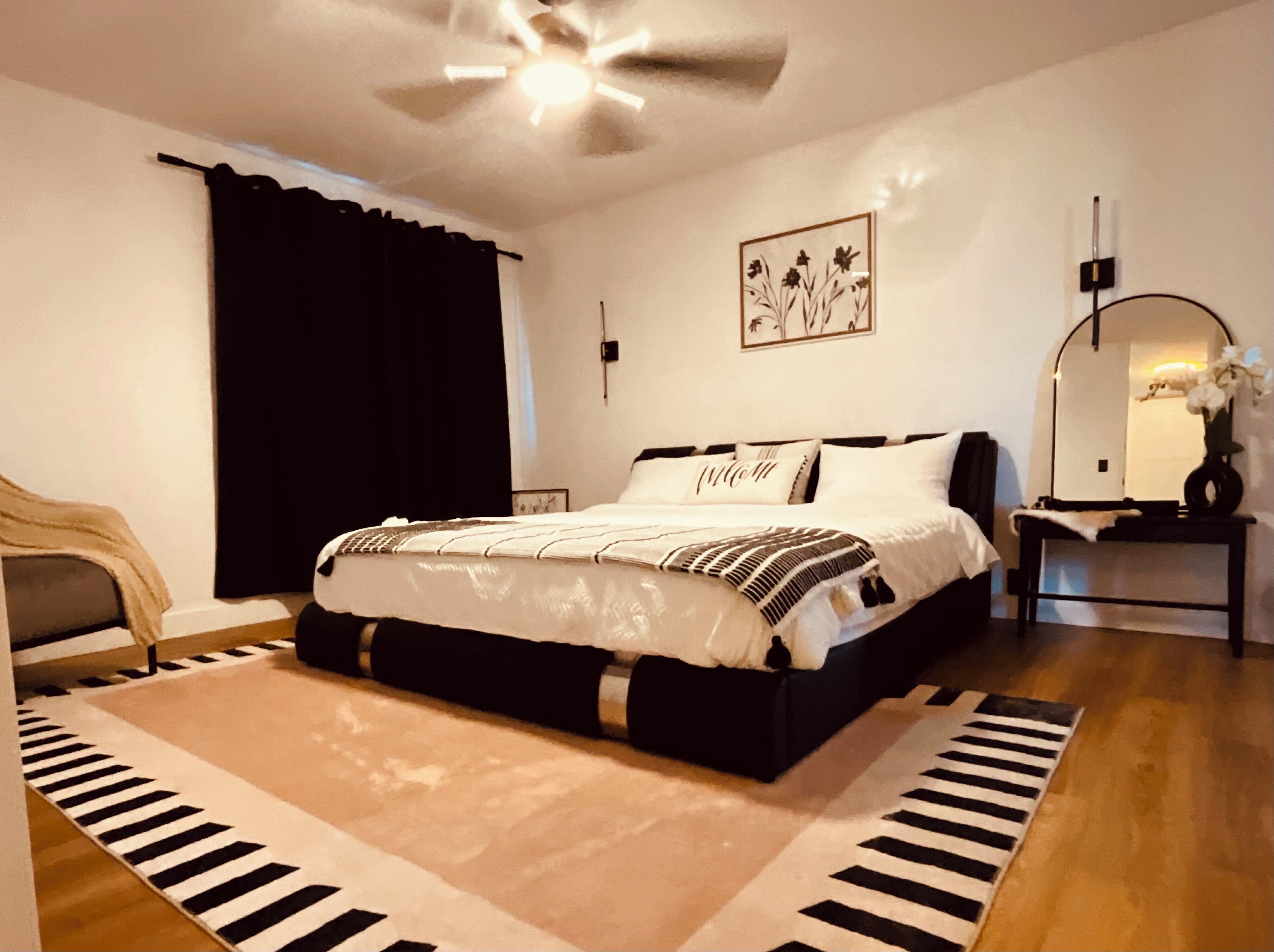 Ein Schlafzimmer mit einem Bett, einem Deckenventilator, einem Nachttisch mit Spiegel, einem schwarz-weißen Teppich und einem schwarzen Vorhang.