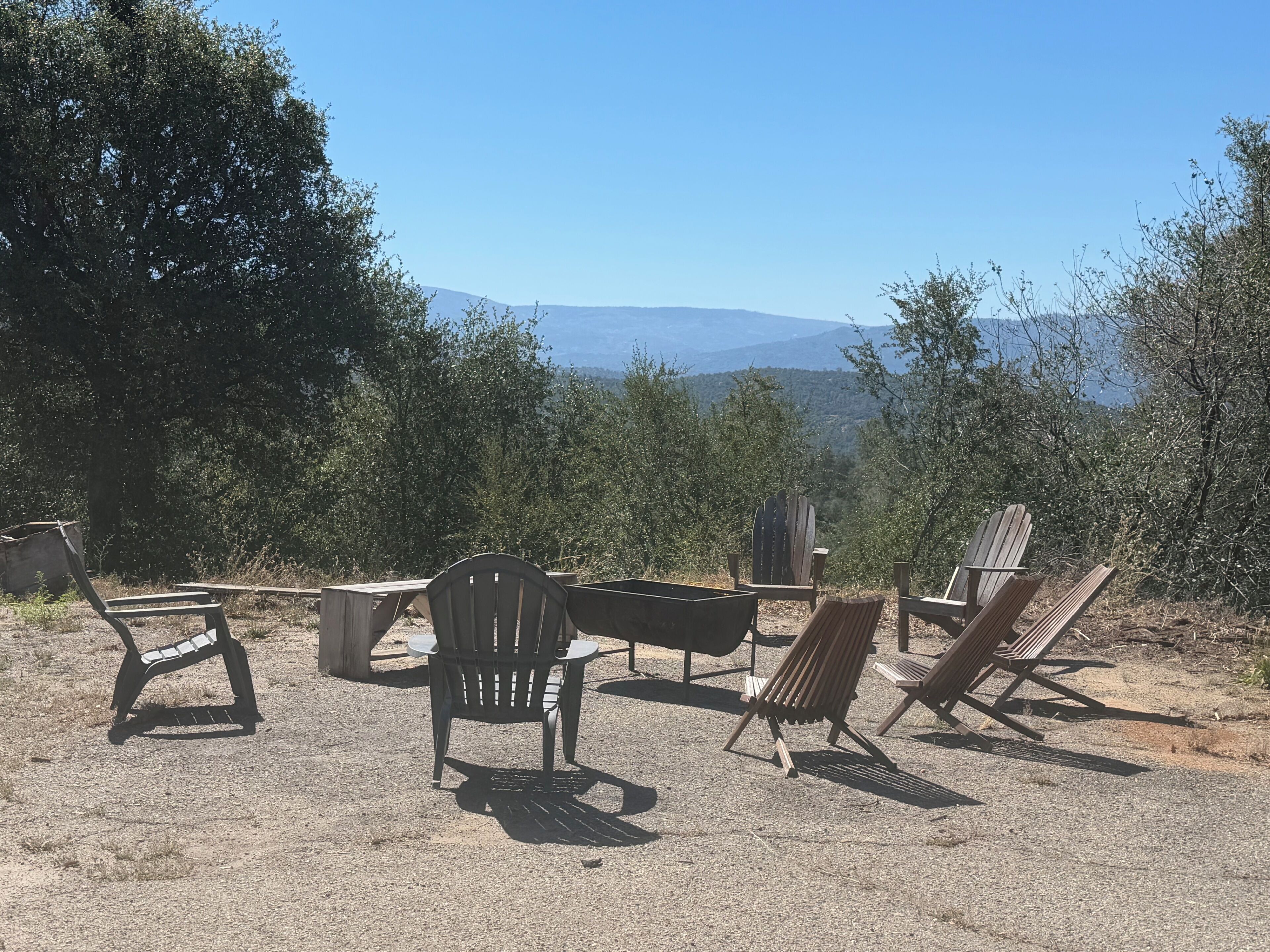 An outdoor seating area with wooden chairs and tables, surrounded by trees and a view of distant mountains.