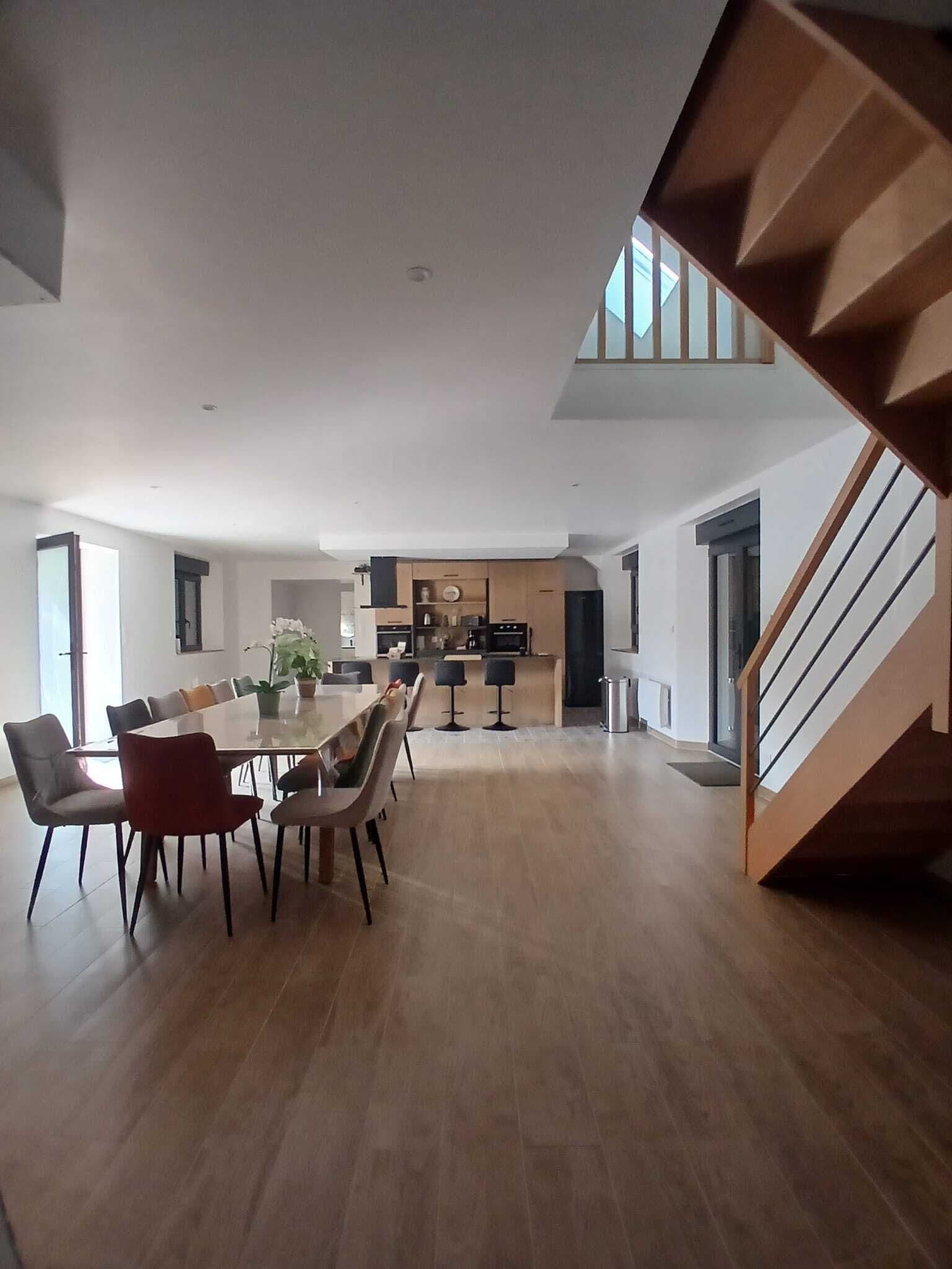 A spacious living area with a dining table and chairs, a kitchen in the background, and a wooden staircase.