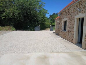 Une allée de gravier mène à un bâtiment en pierre surmonté d’un toit rouge, avec une entrée fermée par un portail et un ciel dégagé.