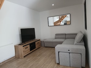 A modern living room with a flat-screen TV, wooden entertainment unit, grey sectional sofa, and a large mirror on the wall.