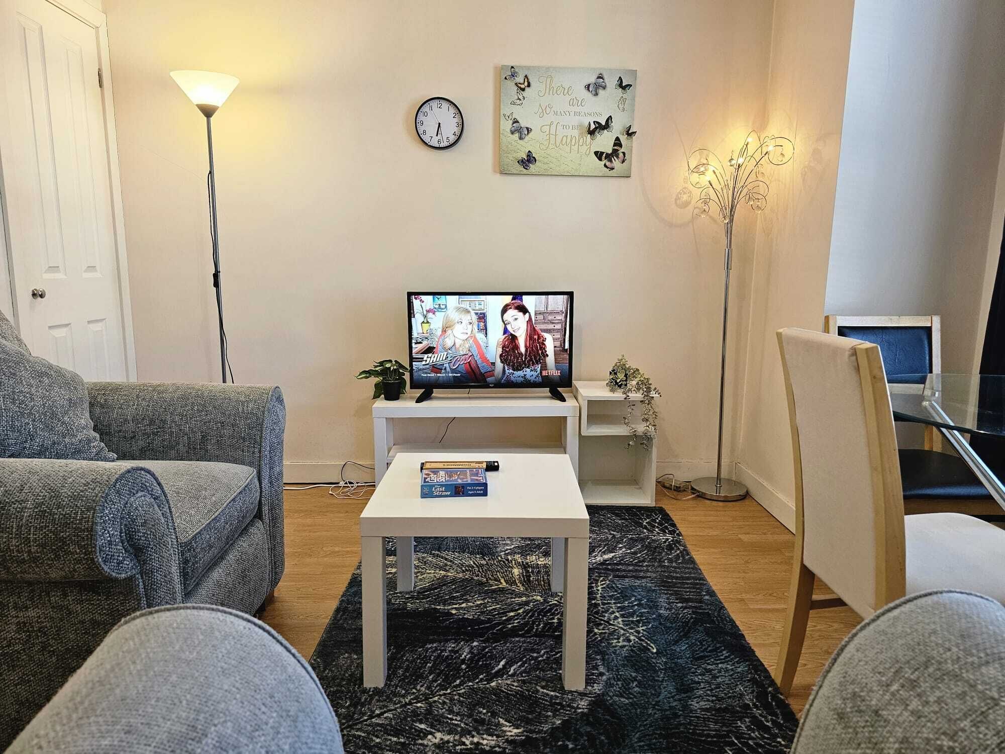Uma sala de estar com televisão, relógio, um letreiro decorativo de borboleta, uma planta em vaso e uma mesa de vidro.