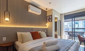 Ein Hotelzimmer mit Bett, Nachttischen, einer Klimaanlage, einer Wandleuchte und Blick auf die Stadt durch große Fenster.
