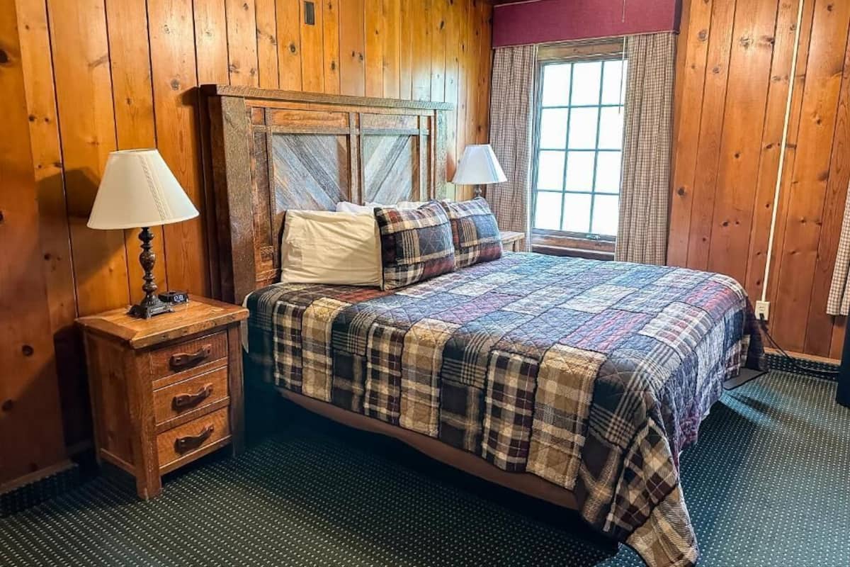 Una habitación con paneles de madera, una cama, dos lámparas de noche, un pequeño mueble auxiliar de madera y una ventana con cortinas.