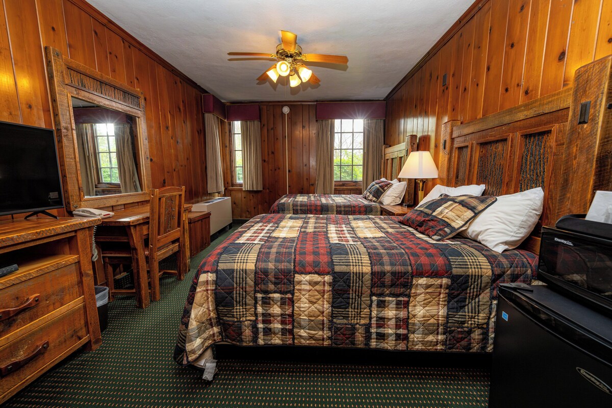 A room with two beds, a desk, a chair, a television, and a ceiling fan.