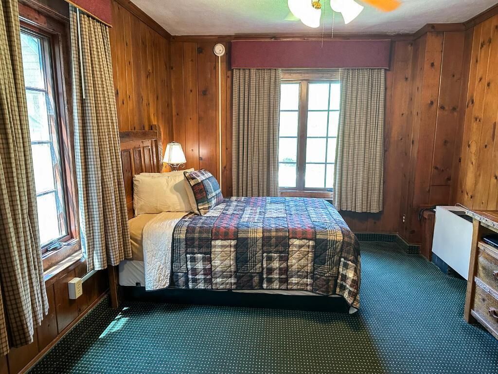 Una habitación de madera con cama, una ventana con cortinas, una lámpara y un radiador.