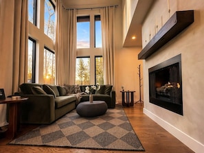 Ein modernes Wohnzimmer mit Ecksofa, rundem Hocker, Kamin und großen Fenstern mit Vorhängen.