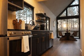A modern kitchen with a stove, oven, and dark cabinetry. There is a window providing a view of trees outside. - Cortland Cabin | Grand Opening (Hendersonville)