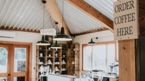 Interior de una cafetería con barra de café, luces colgantes y estantes con artículos.