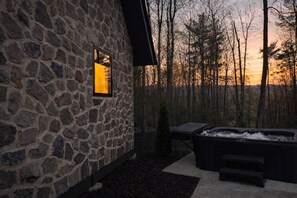 A stone building with a window, a hot tub, and a view of trees at sunset. - Cortland Cabin | Grand Opening (Hendersonville)
