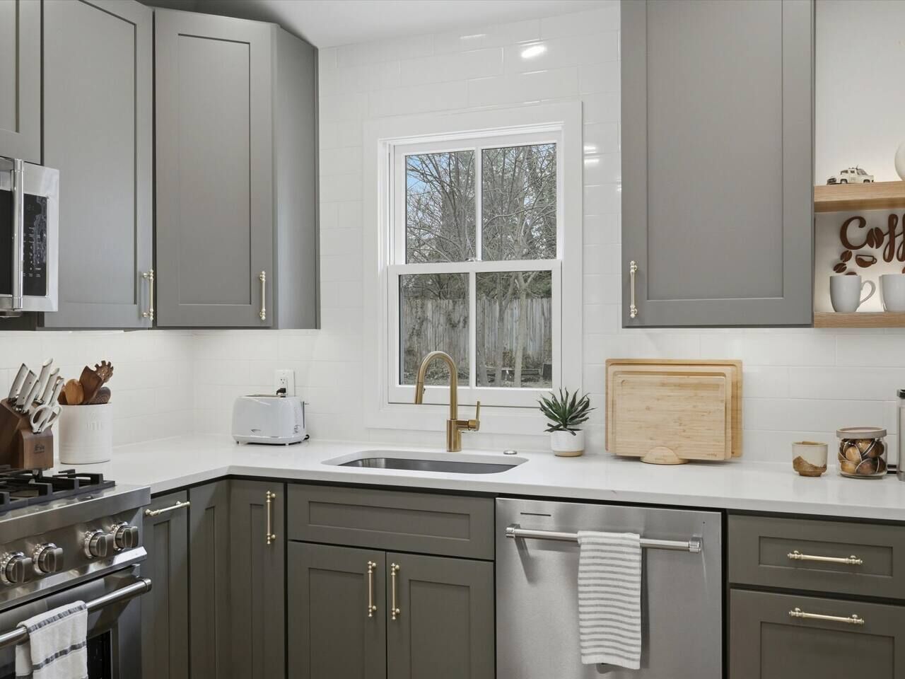 A modern kitchen with grey cabinets, a white countertop, a stainless steel sink, a gas stove, and a window with a view of trees.