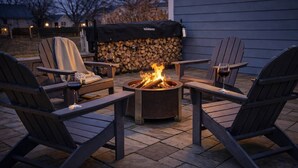 Un foyer entouré de fauteuils Adirondack, un barbecue couvert et un mur de pierres avec des bûches.