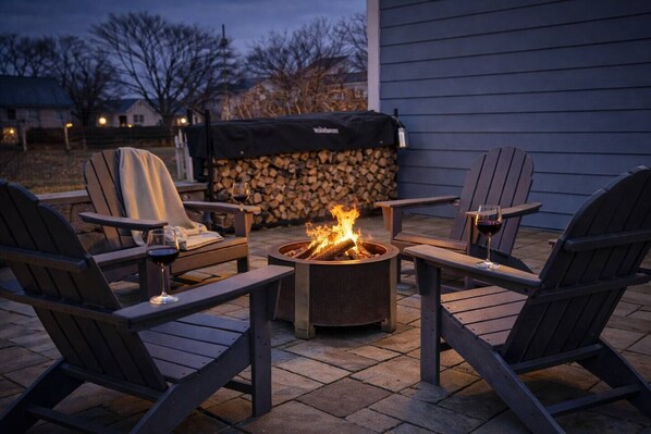 A fire pit surrounded by Adirondack chairs, a covered grill, and a stone wall with logs.